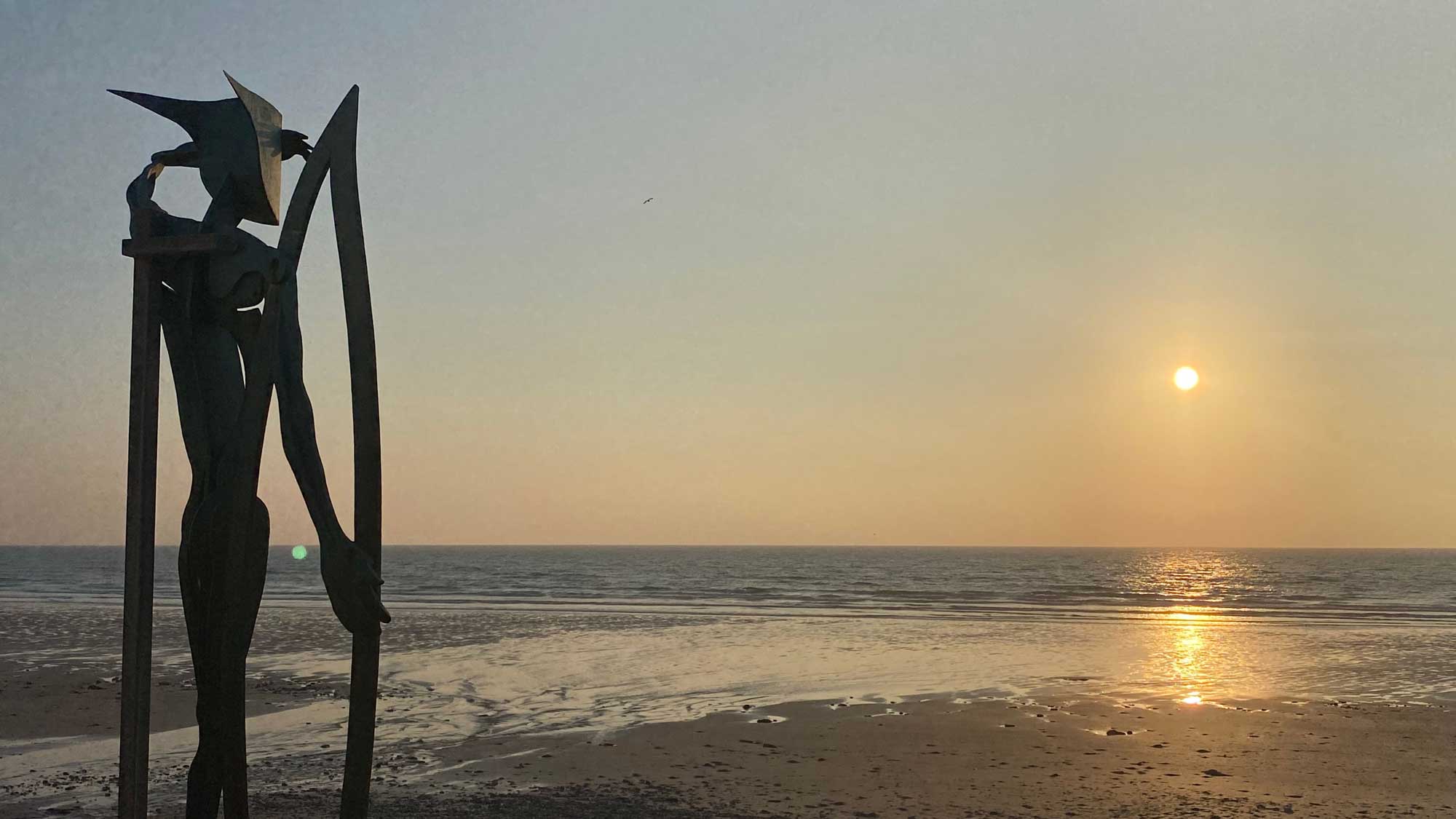 a sculpture of a figure looking out to sea as the sun sets over the bay