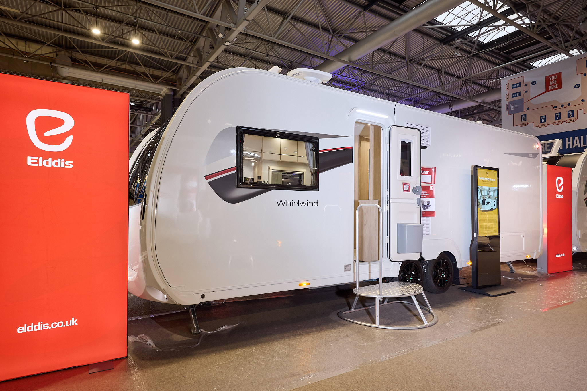 The Elddis Whirlwind 860 has a white exterior with a grey, red and black decal. The entrance door is open and there is a metal step to gain easy access. There is a large Elddis red banner with their website address next to it.