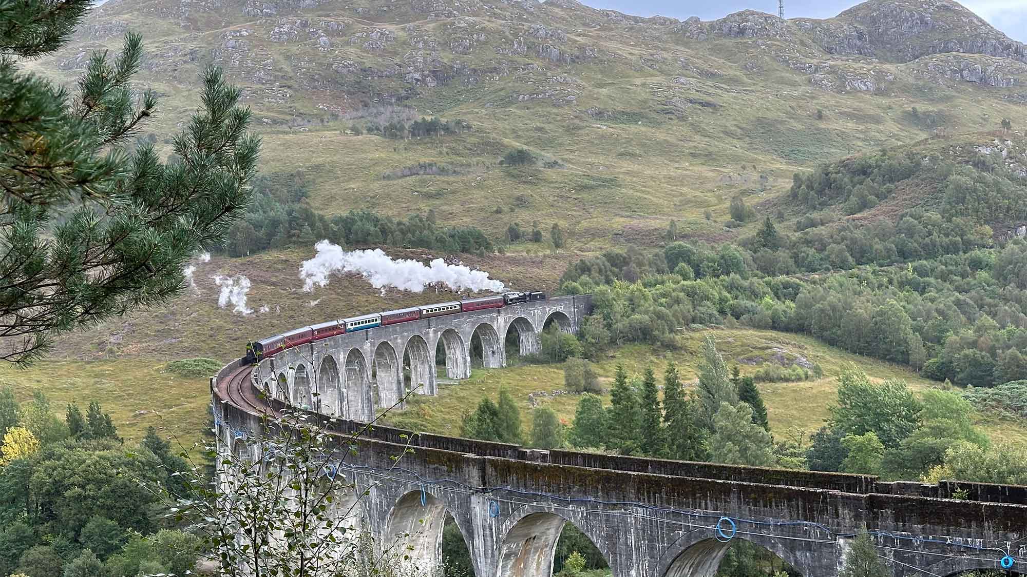 /a tall viaduct curving around with the mountains behind, the steam from the train seen rising into the sky