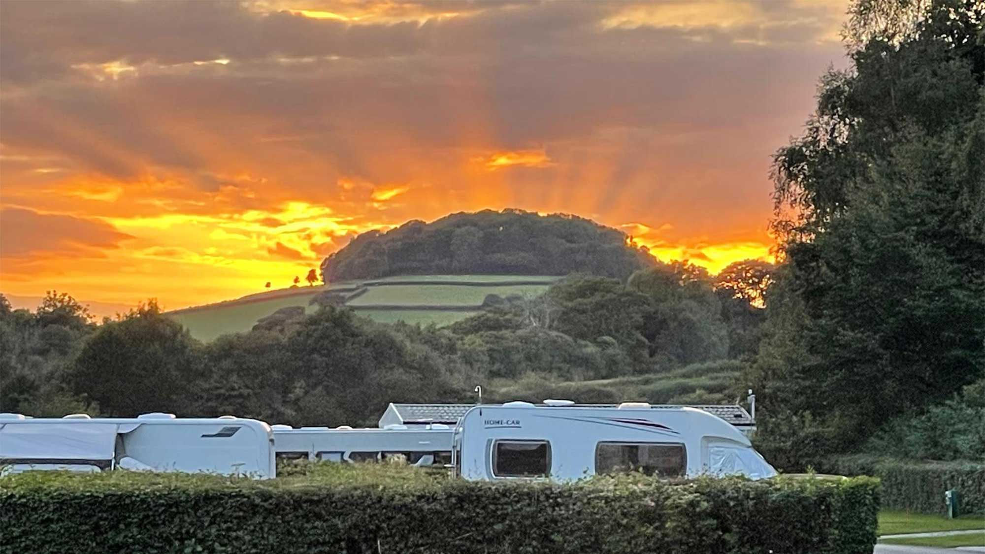 Caravans peeking over a hedge with a beautiful orange sky with the sun setting behind a hill in the distance
