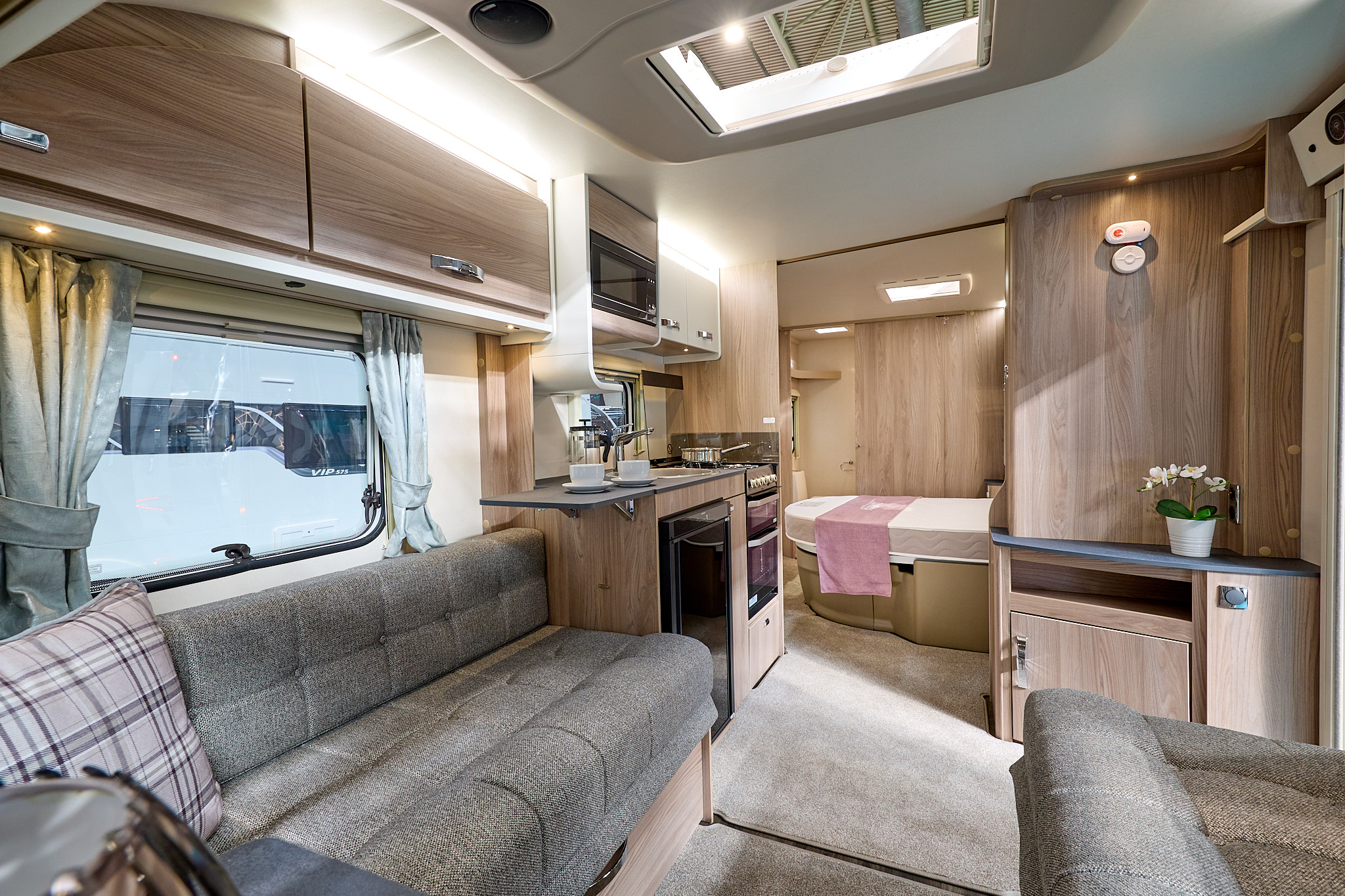 Swift Sprite Major 4 EB's interior has wooden furniture with beige upholstery and carpet.  It has dark grey work surface.  There is a pink cushion and runner on the bed.  The bedroom is to the rear.  The skylight is open.