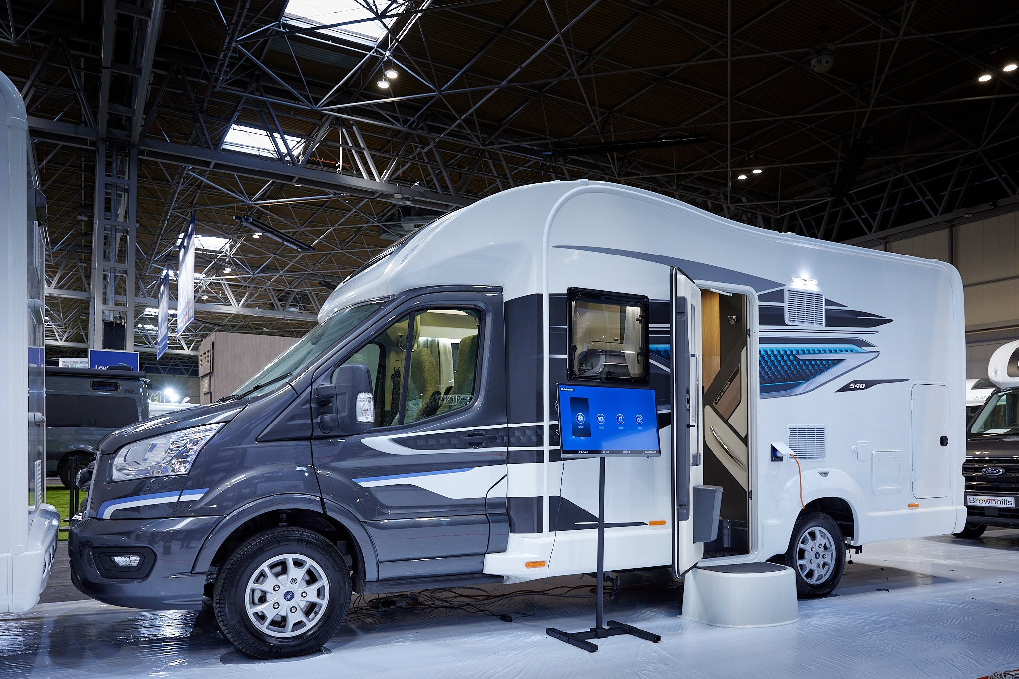 Swift Voyager 540, the vehicle has a grey cab and a predominantly white body, its habitation door is open showing into the interior, with a step to gain easy access.  There is an interactive screen outside the Voyager..