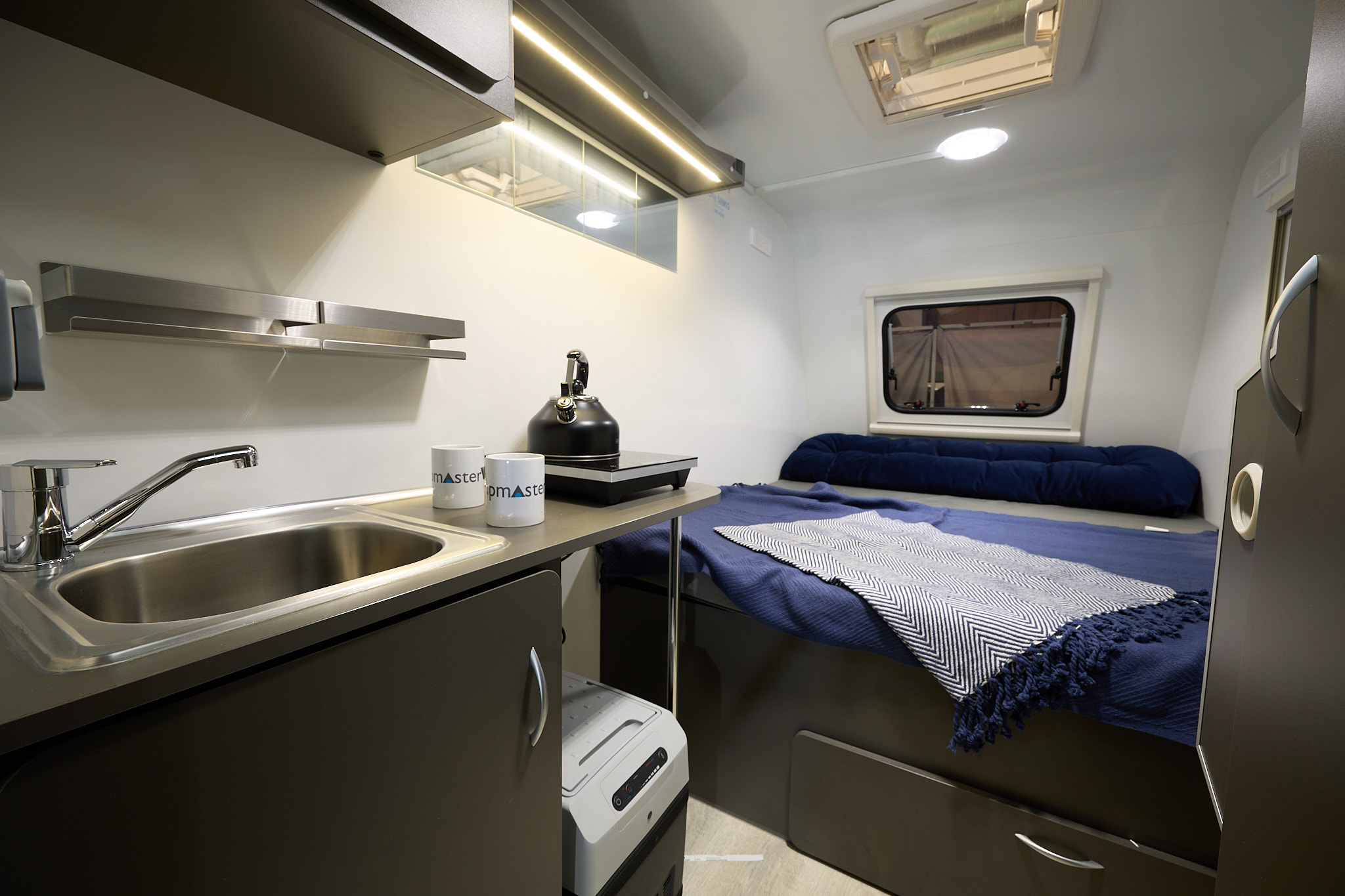 Campmaster Duo GLX interior, kitchen sink and mixer tap, with two mug and a kettle, fixed double bed with blue duvet cover