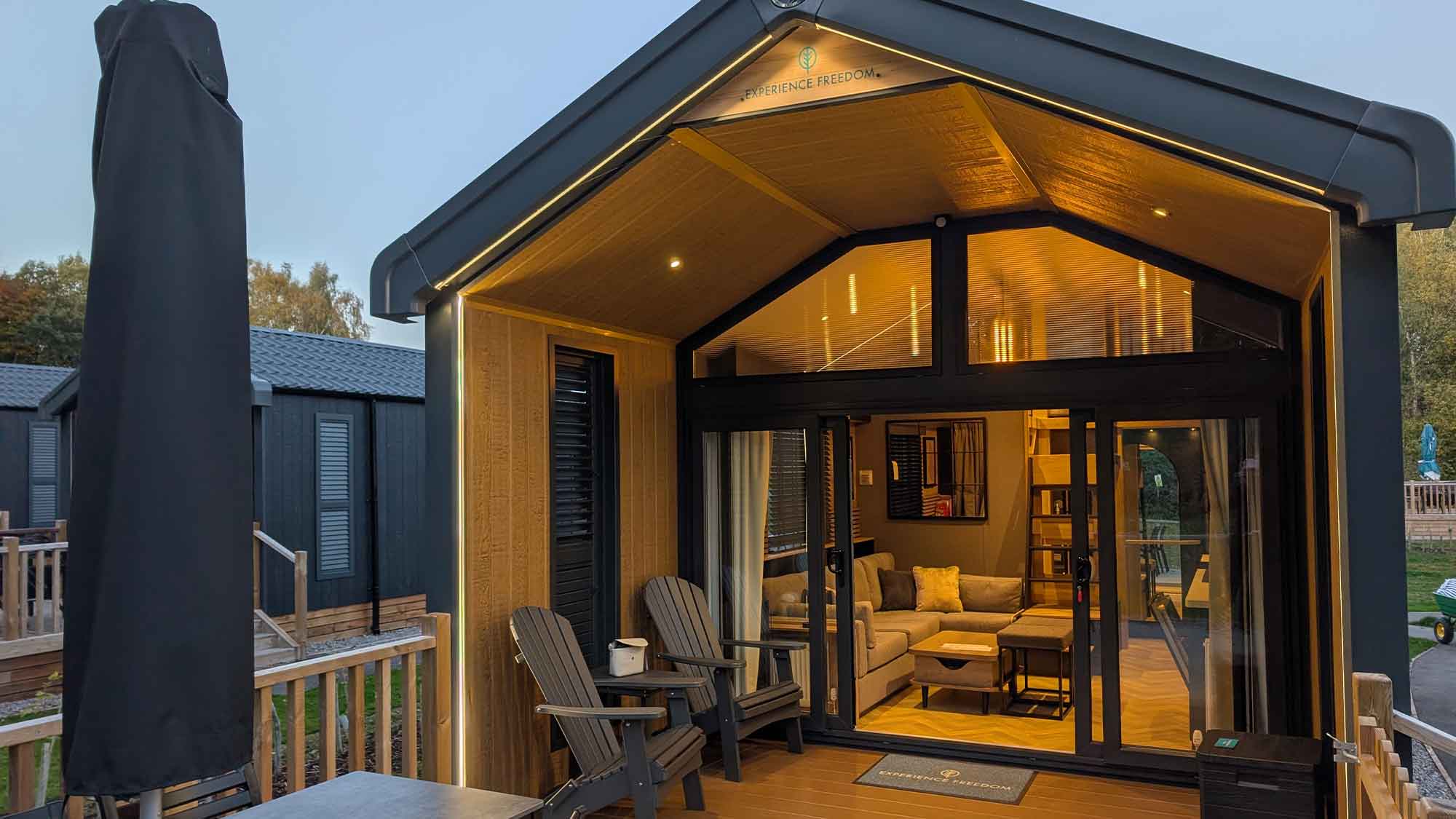 An image of a deluxe glamping cabin from the outside. There are seats outside on the porch, which leads to a glass front. Inside the light is glowing amber making it look cosy and welcoming.