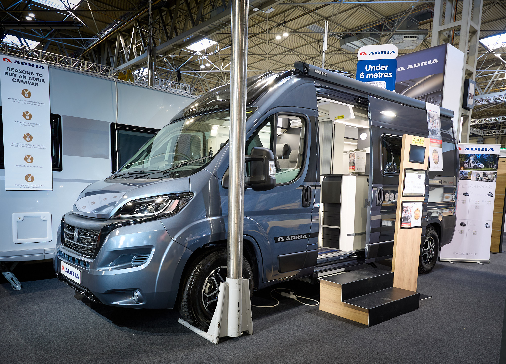 Adria Twin Supreme's exterior is metallic blue, the sliding door is open and there are wooden steps to gain easy access.  There is an interactive information board by the side of the van.