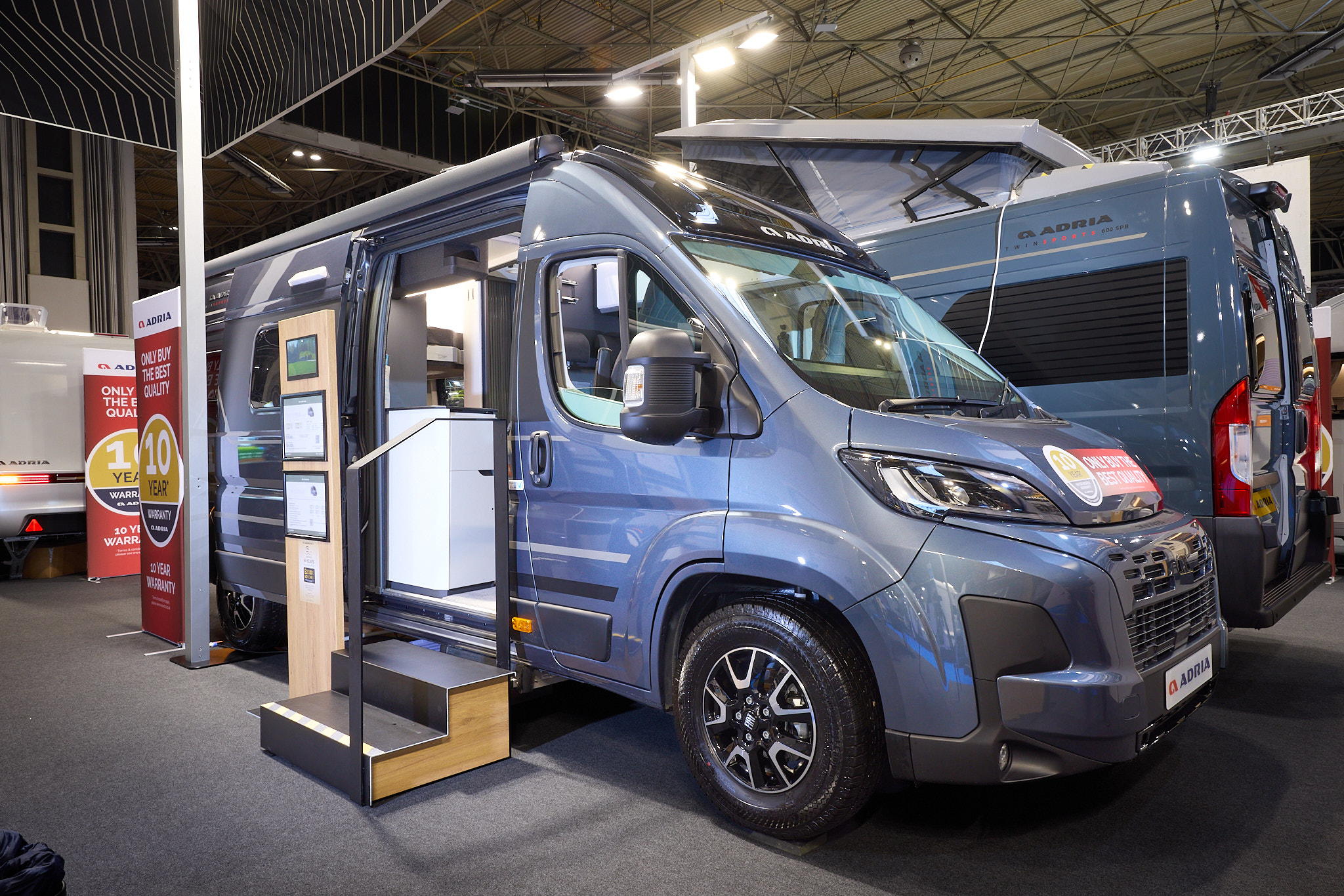 The Adria Twin Supreme has a blue exterior.  The habitation door is open and there are two steps to gain easy access. There is an information board to the left of the steps.  