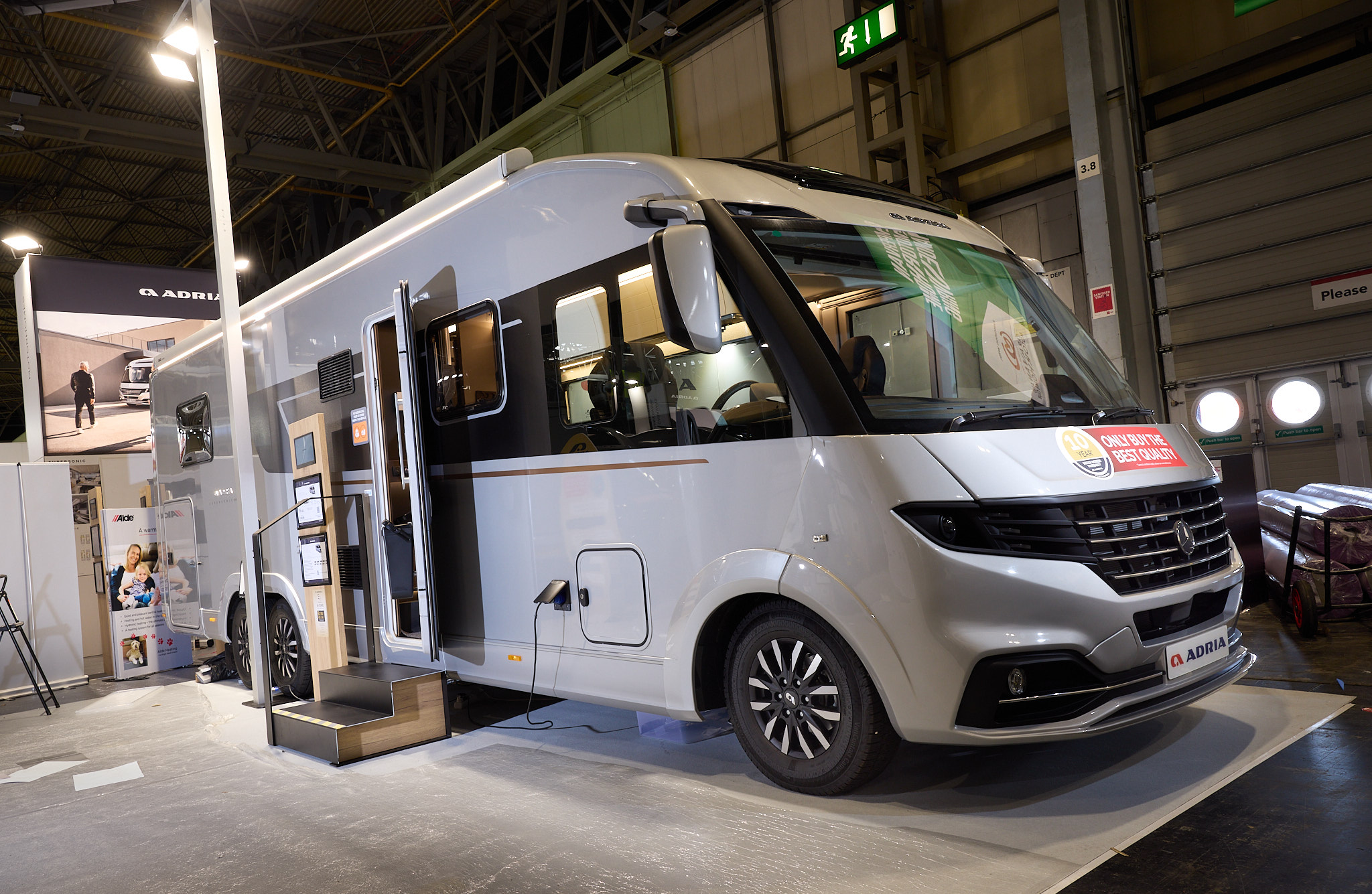 The Adria Supersonic has a silver exterior.  The habitation door is open and there are two steps to gain easy access. There is an information board to the left of the steps.  