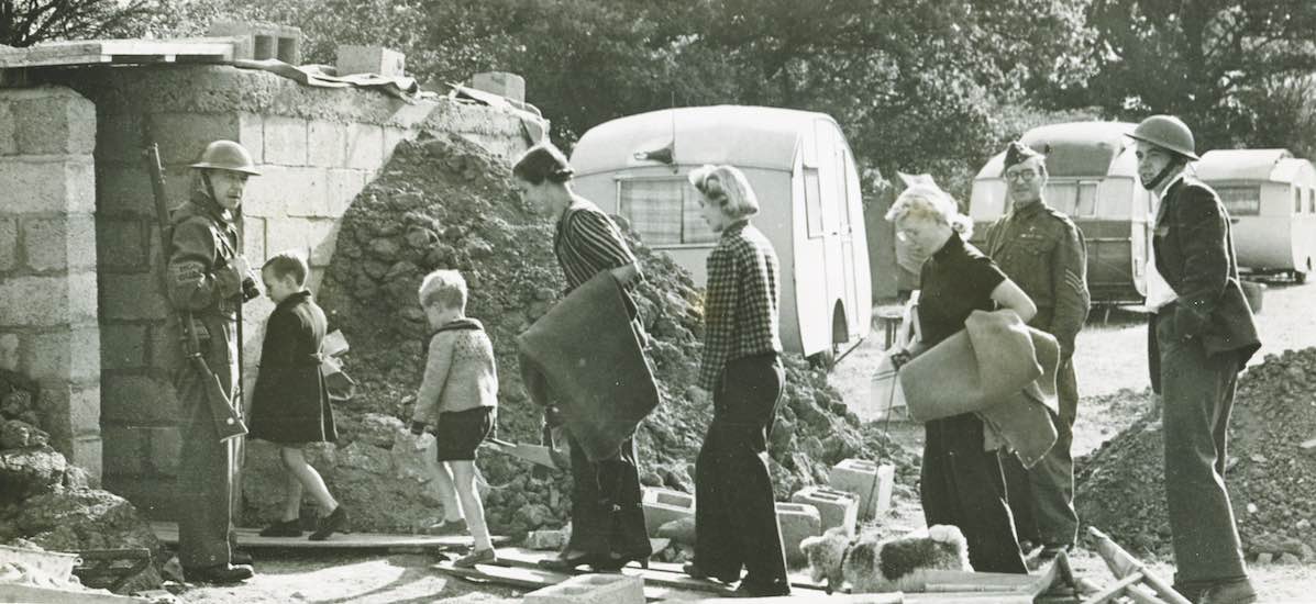 Citizens walk on duckboards past rubble with Home Guard soldiers looking on and caravans in the background