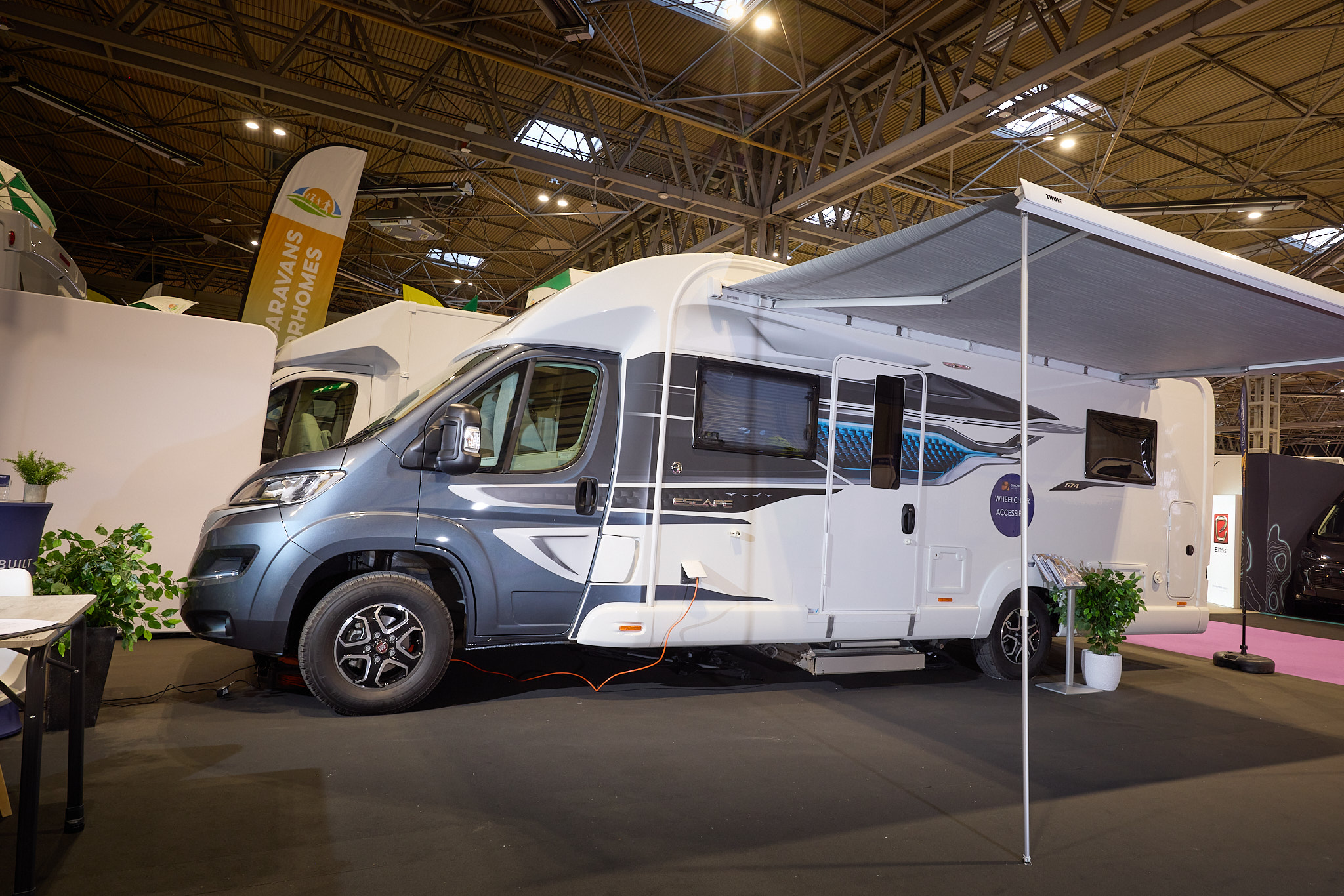 The Coachbuilt GB LIV Swift Escape 674 has a great cab with white body.  There are grey and turquoise decals.  There is a large canopy awning with a stand and plant to the right of the habitation door.