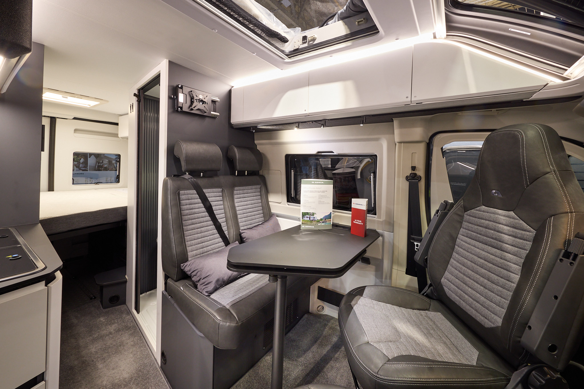 The Adria Twin Sport’s floor has a mid brown carpet, with white cabinets and dark grey panels.  Its rising roof is open.  The upholstery is two tone with dark edging.  There is a grey table which can be rotated.  