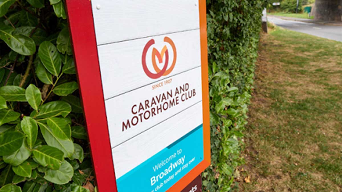 Braodway Club Campsite sign board at the entrance with a green leafy hedge behind and grass along the side