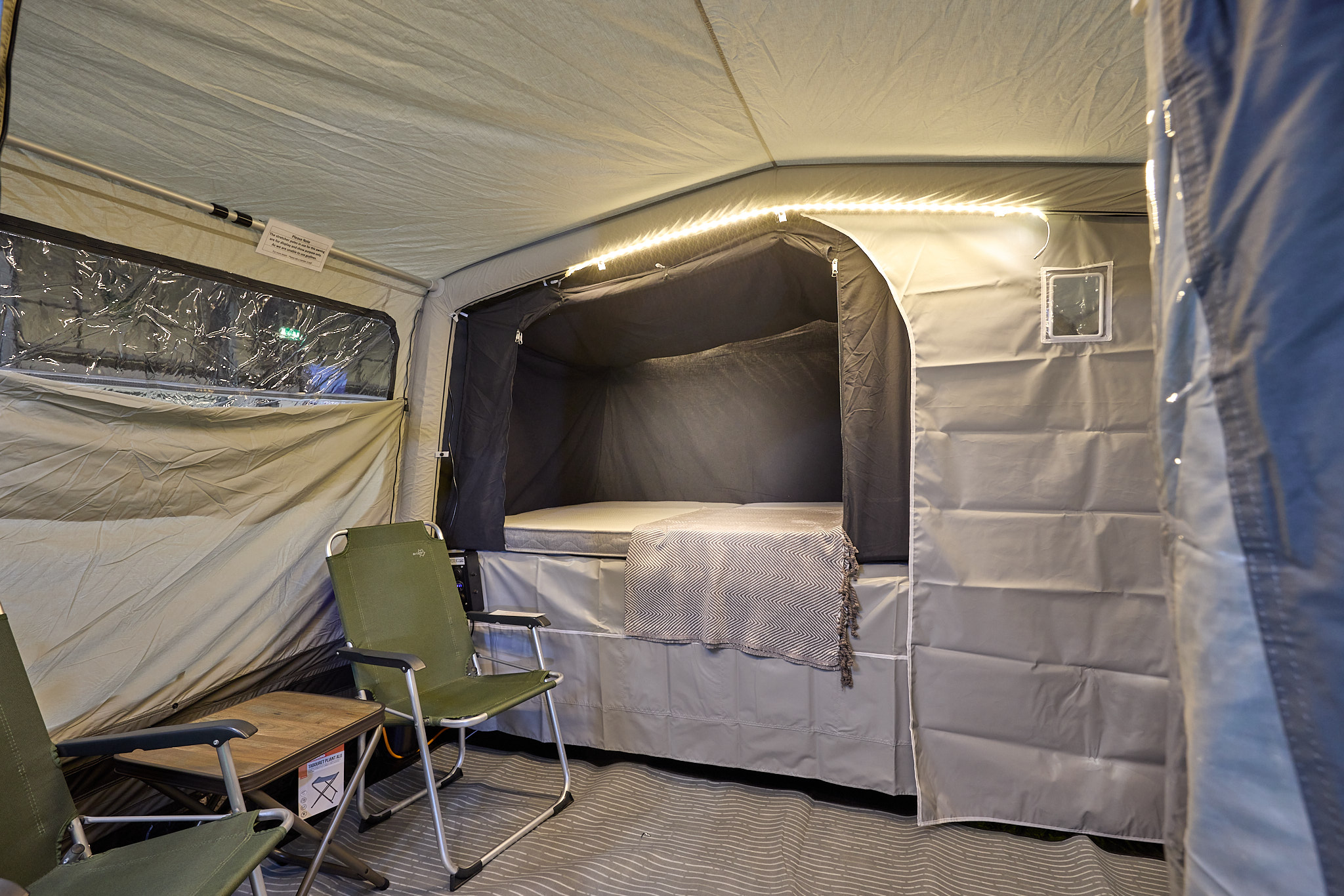 In the Campmaster 1000 LX there is a coffe table and chairs.  The bedroom door is open. There is LED strip lighting on the ceiling.