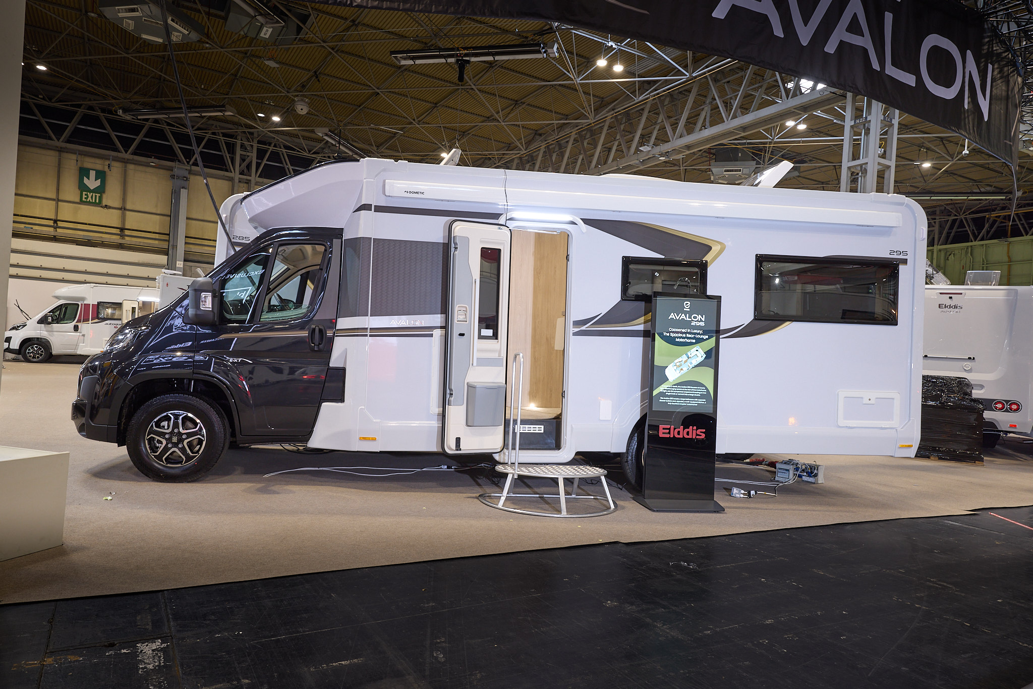 The Elddis Avalon 295 has a black cab and a white body with black and gold decals.  There is a metal step by the open habitation door, revealing the interior, there is an interactive stand to the right.