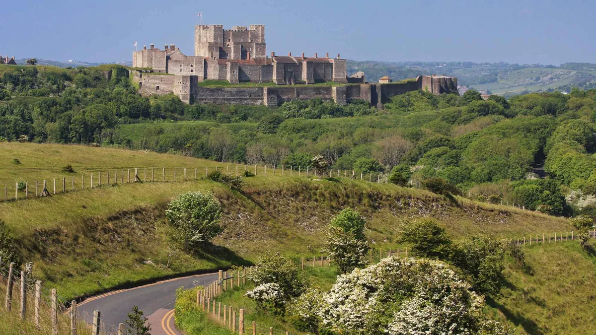 A slightly elevated view looking across at Dover Castle in the distance with a hazy sky beyond. A windy road is just below leading off beyond grassy banks and green trees. 