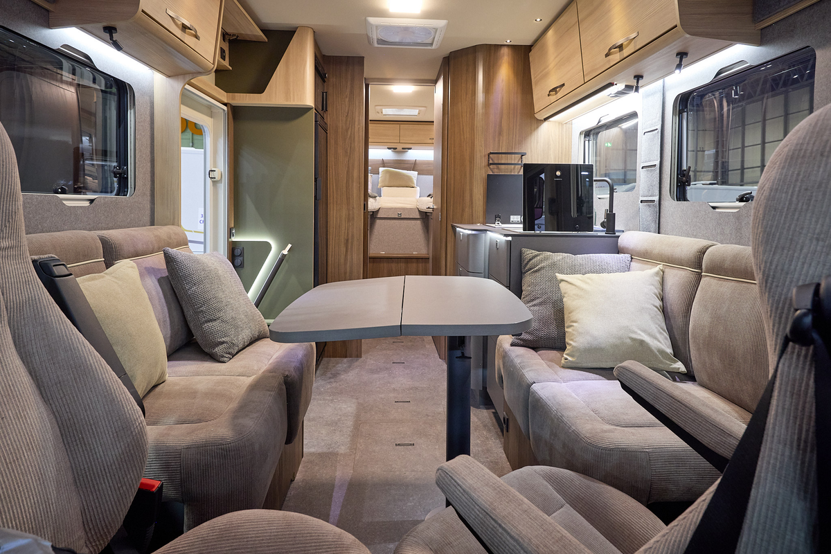 The Eura Mobil Integra Line GT 726 EF has beige carpets and wooden cabinets.  The upholstery is mid brown.  There is a grey table (extended) in front of the sofas.  There are two steps to the rear fixed bed.