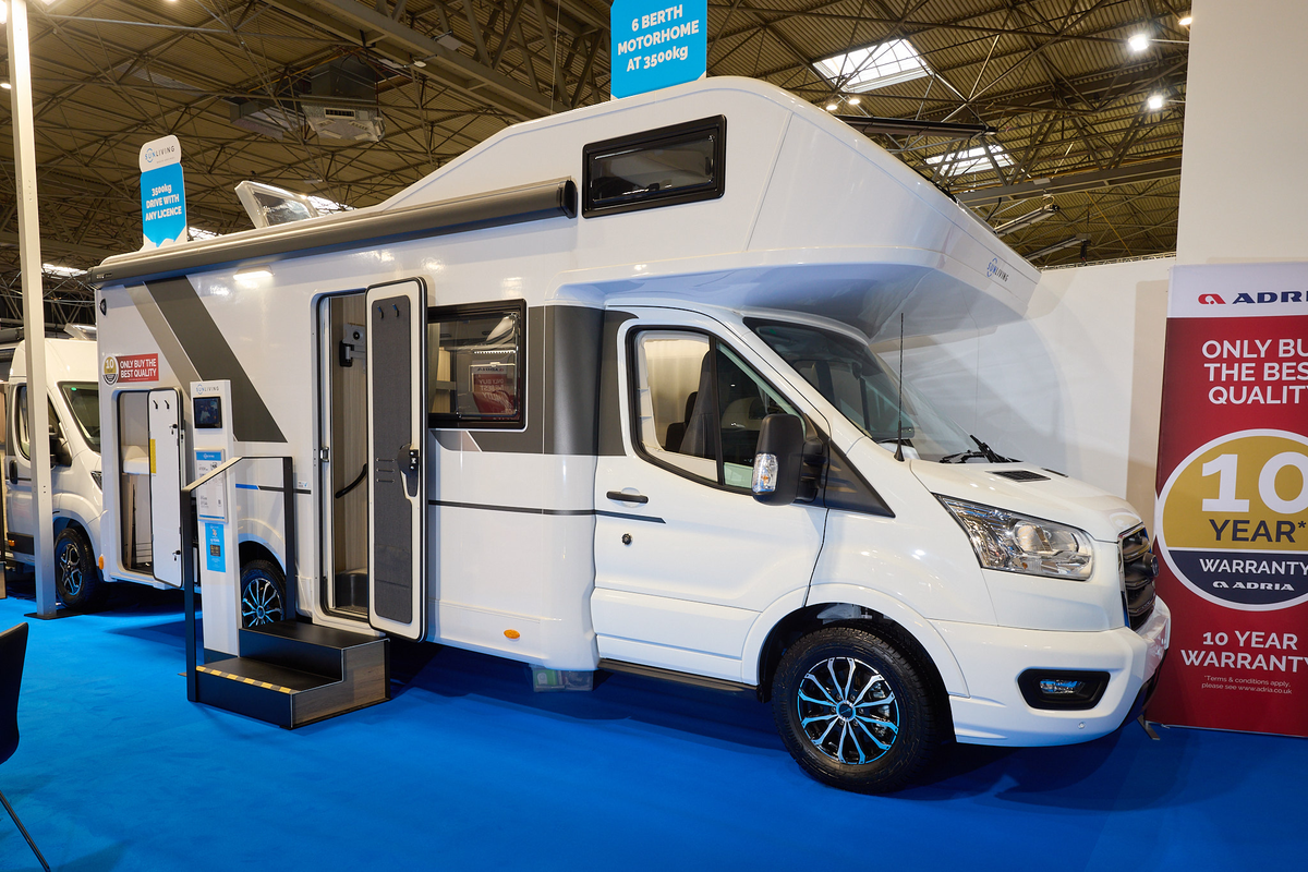 The Sun Living A Series 70DK is white with brown and bronze decals.  There are two steps outside the open habitation door.  There is an interactive stand next to the steps and the rear locker door is open.