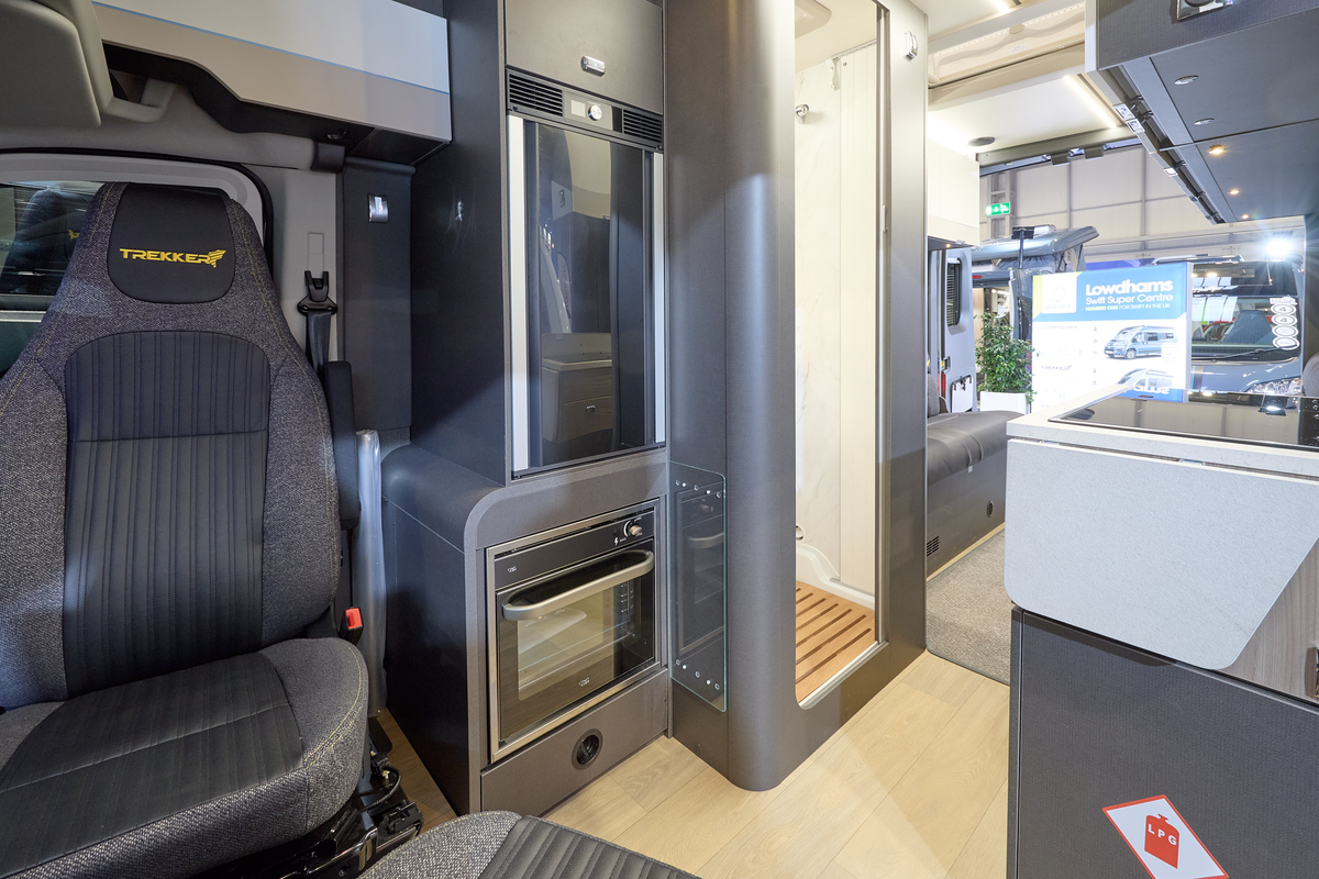 The Swift Trekker XL has a wooden floor with a step up into the grey carpet at the rear.  There is an oven behind the driver's seat.  The washroom door is open.  There is a rear lounge.