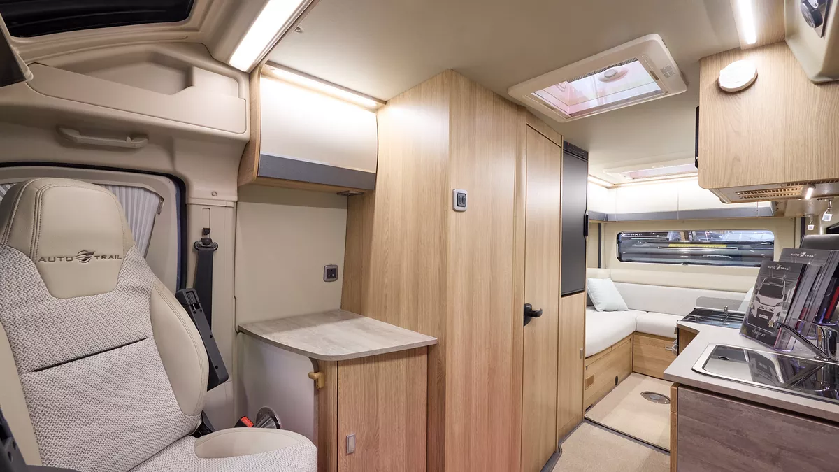 The Auto-Trail V-Line 635 Sport has beige carpets, with pale wood cabinets with cream and grey overhead lockers.  The upholstery is white and cream.  There is a rear lounge.  The front seats are swivelled around.  There is a roof light.