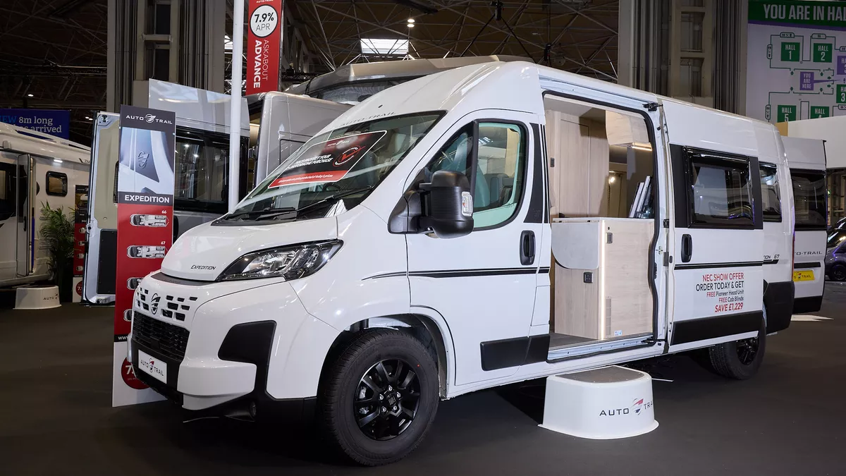 The Auto-Trail Expedition 67 is white with black decals.  There is a white step by the open sliding door which reveals the interior.  Its rear doors are open.