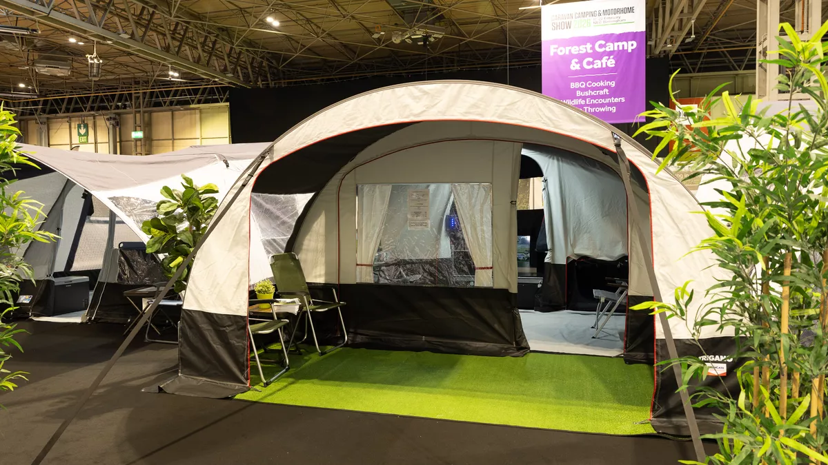The Trigano Nebra has cream and brown canvas.  It has a curved structure.  There are two green chairs in the awning area which has a faux grass mat.  The entrance door to the living area is open.