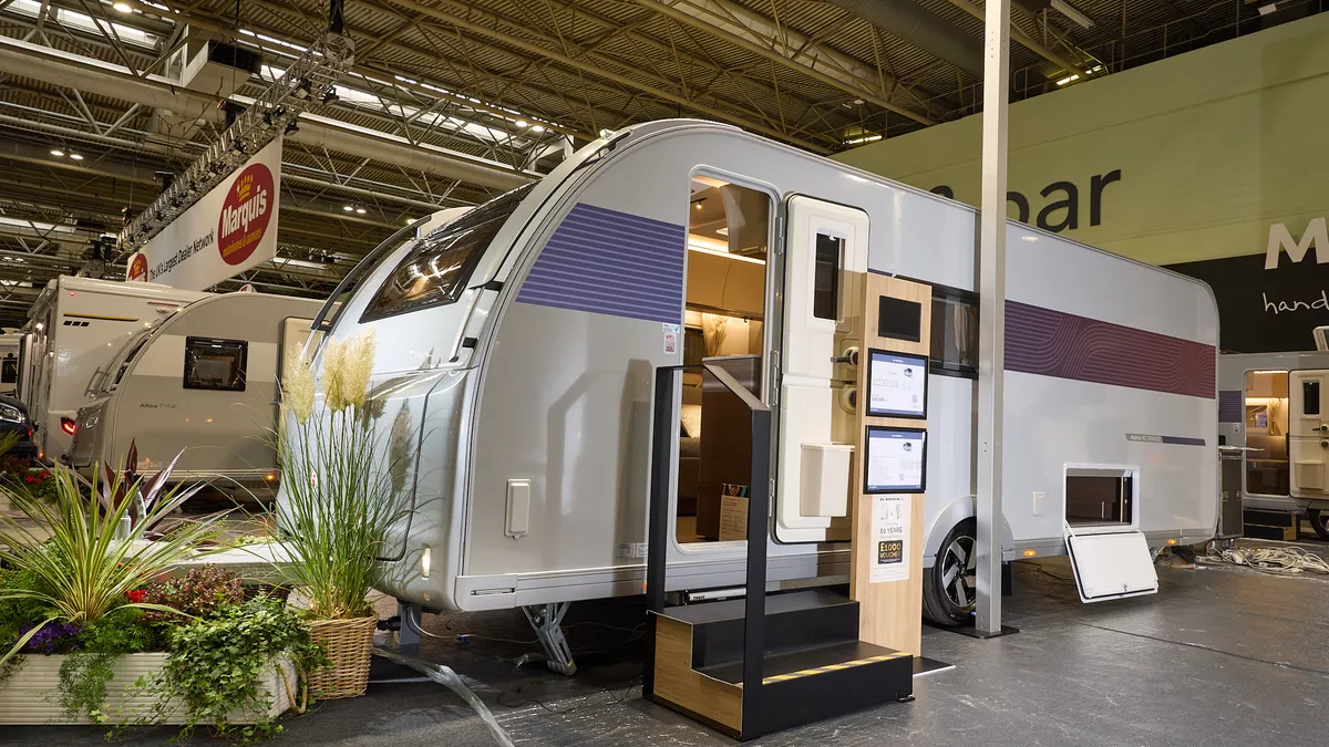 The Adria Alpina Rio Grande has a silver exterior with a blue decal that turns to purple at the rear, its entrance door is open and there are two wooden steps to gain easy access.