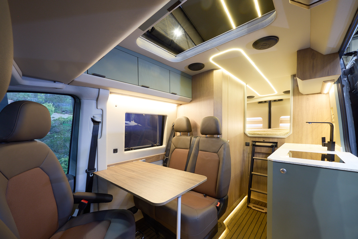 The Kaktus Adventure Vans Dune x4 has a wooden floor.  The upholstery is two tone brown leather.  The cabinets are grey.  There is a ladder to the fixed rear bed.  The pop top roof is extended.