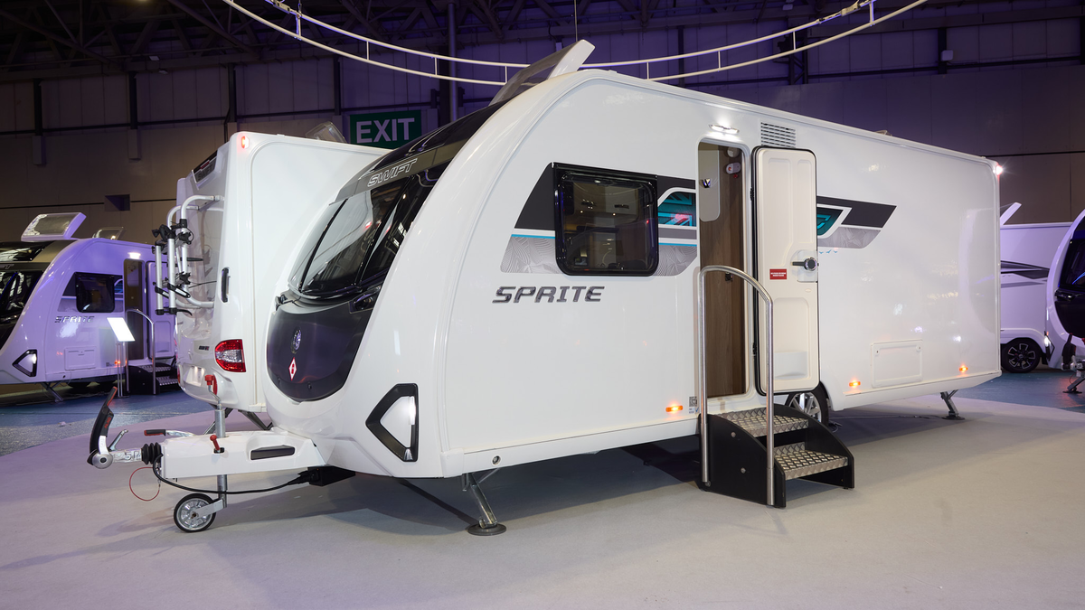 Swift Sprite Major 4 SB’s exterior's white with a black panelled front.  There's a turquoise decal to the right of the side window with the Union flag.  The entrance door is open and there are two metal steps to gain easy access.  The roof light is o