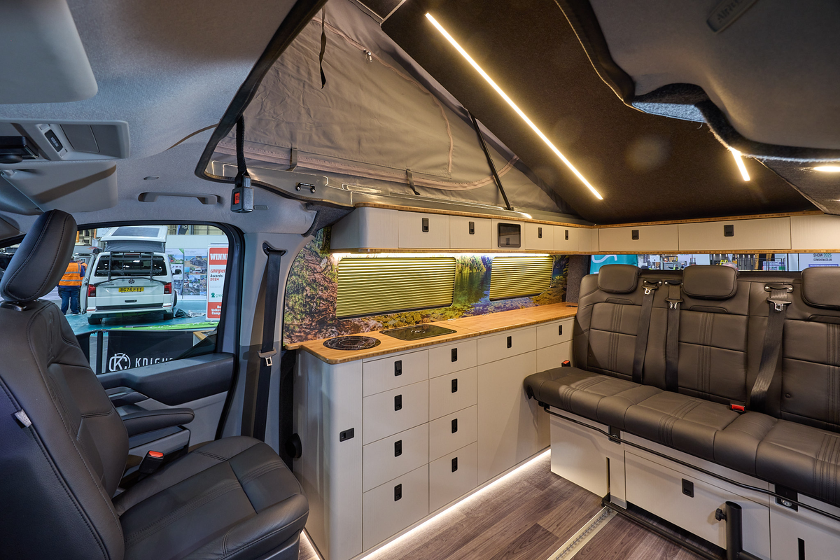 Knights Custom Conversions Prestige Tourer Plus has wooden flooring with black upholstery.  The cabinets are white and pale wood.  The pop top roof is open.