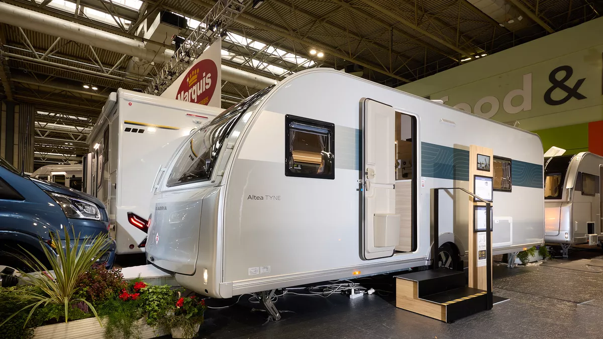 The Adria Altea Tyne has a silver exterior with a blue decal that fades towards the front, its entrance door is open and there are two wooden steps to gain easy access.  