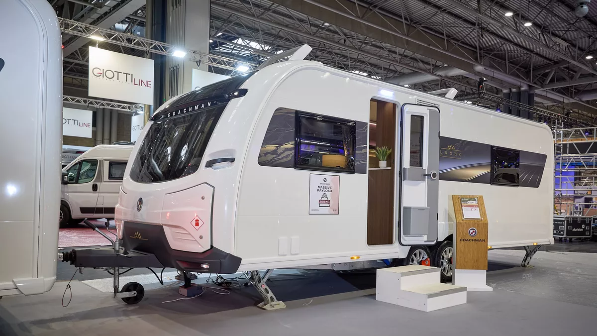 The Coachman LUSSO II has a white exterior with a brown patterned decal.  Its sunroof is open.  The entrance door is open and there are two wooden steps to gain easy access.  There is a stand next to the steps.