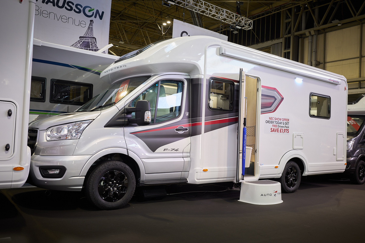 The Auto-Trail F-Line F74 has a white body with grey and red decals.  There is a show offer on its side in red.  There is a white step by the opened habitation door.  