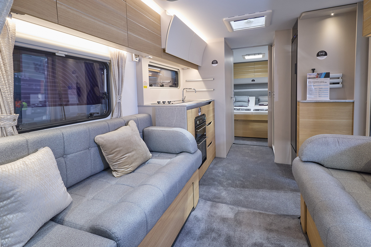 The interior of the Adria Adora 623 DP Tiber has cream walls.  The furniture is mid wood.  There are two sofas with grey upholstery and on one there are two cushions.  There is a large fixed bed at the rear.