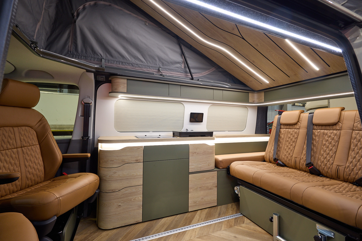 The Sherwood Campers Ltd Loxley Edition Conversion has wooden parquet-style flooring and its upholstery is brown leather.  Its furniture is wooden which is curved.  There are dark green inserts.  The rising roof is open, there's strip lighting.