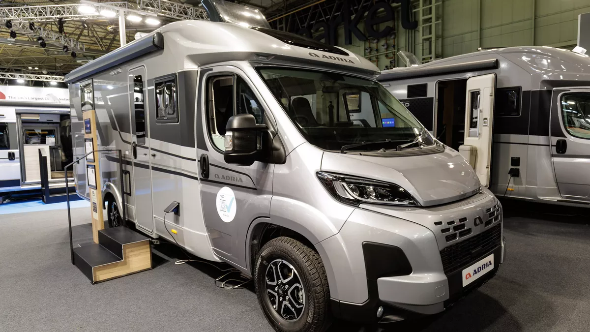 The Adria Compact Supreme DL is silver with a white coachbuilt body.  It has dark grey decals along its side.  There are two steps up to the main door to enable easy access.  It has an interactive stand by the side of the steps.