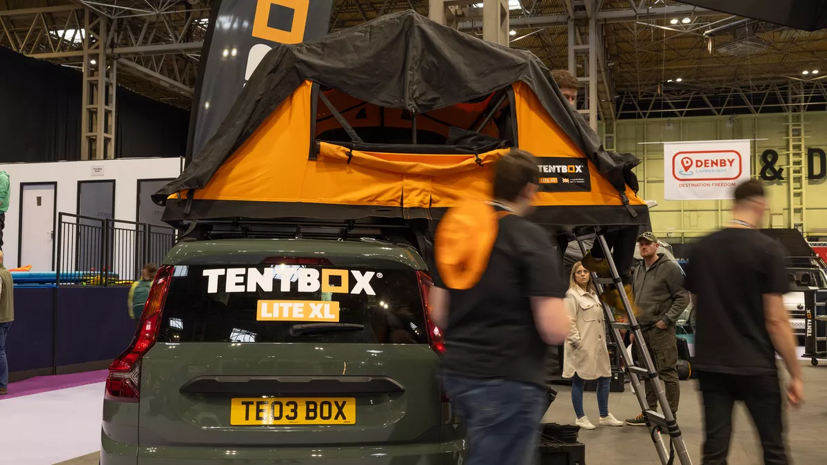 The TentBox Lite XL has orange and black canvas, its window is open. There is a silver ladder to gain access.  It is displayed on a green car.