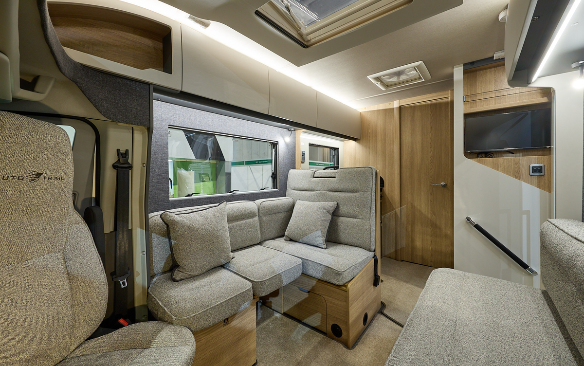 Auto-Trail Excel 675B’s interior has beige fabric upholstery with matching cushions.  The furniture is wooden.  There is a TV above the handrail by the entrance door.  There is one sky light.  The internal door to the washroom is closed.