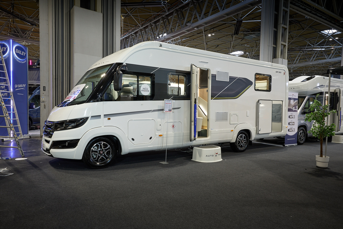 Auto-Trail Grande Frontier GF-80's exterior is white with dark blue and black decals.  The entrance door is open revealing a handrail and there is a white step to gain easy access.  There is a roller banner to the rear.