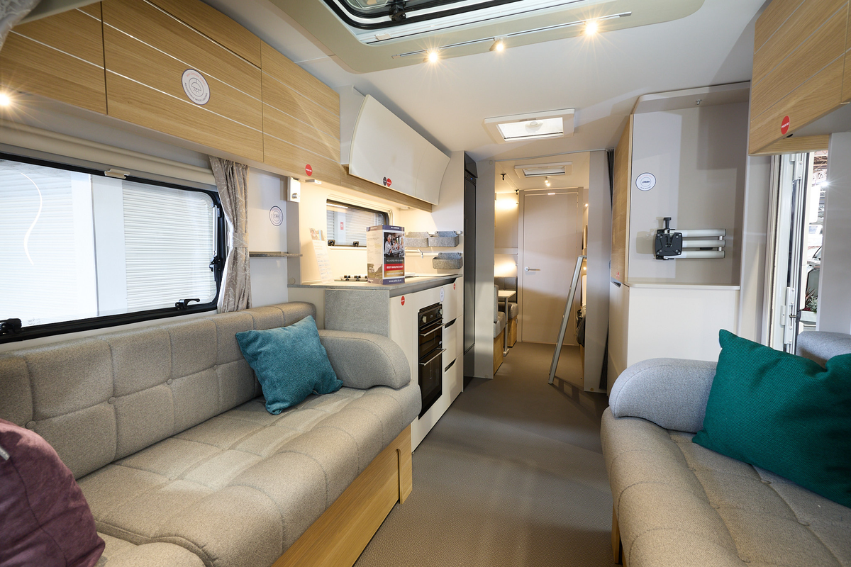 Adria Adora Sava’s interior, it has wooden cabinets with grey upholstery in the lounge.  There are three cushions, blue, green and purple.  The kitchen has a domestic style oven and hob.  There is a ladder to the top bunk.
