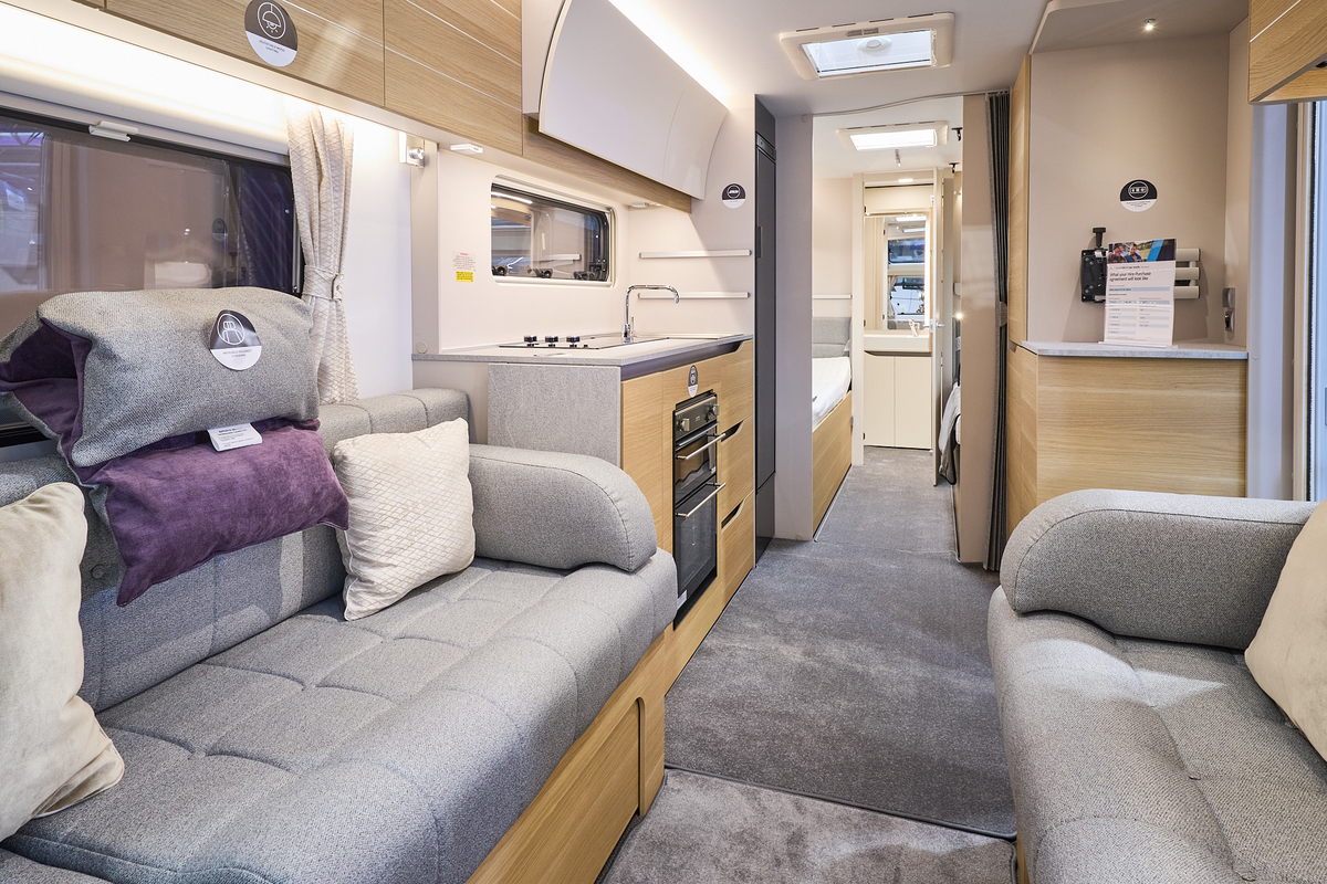The interior of the Adria Adora 612 DL Seine has cream walls.  The furniture is mid wood.  There are two sofas with grey upholstery and on one there is a head rest consisting of a grey and a purple cushion.  The washroom is at the rear.