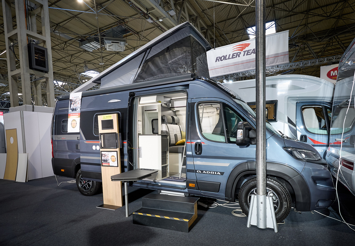 Adria Twin Sports’ exterior is metallic blue, the sliding door is open and there are wooden steps to gain easy access.  There is an interactive information board by the side of the van.  The rising roof is extended.