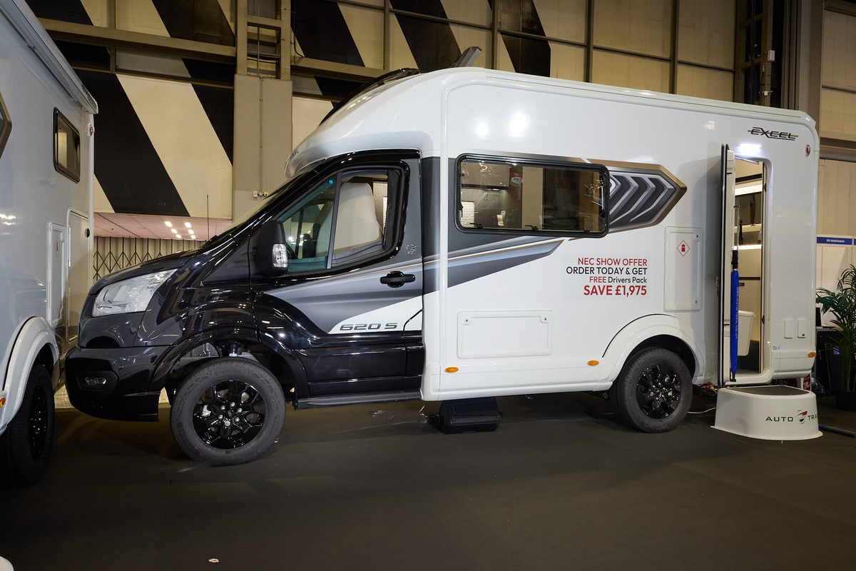 The Auto-Trail Excel 620S has a black cab and white body with grey decals.  There is a show offer on its side in red.  There is a white step by the opened habitation door which reveals the rear kitchen.