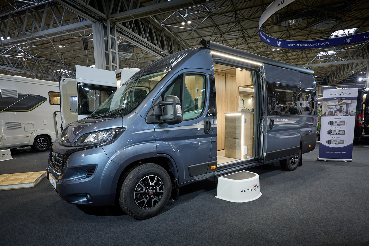 Auto-Trail V-Line 635 Sport's exterior is mid grey with silver decals.  The sliding door is open revealing the kitchen and there is a white step to gain easy access.  There is a roller banner to the rear.
