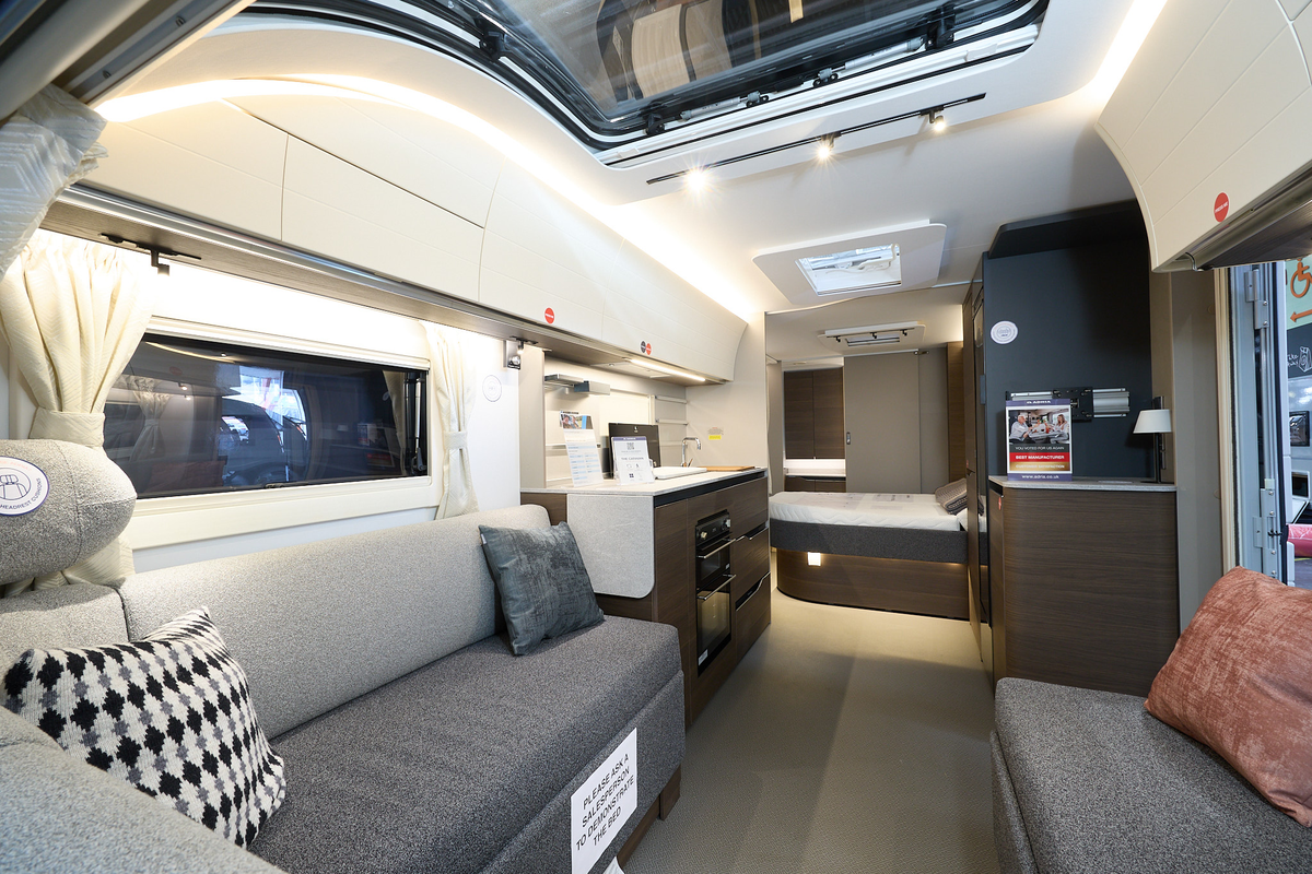 Adria Alpina Mississippi’s interior, it’s cream with a large panoramic window.  The upholstery is two tone grey.  The fixed bed is beyond the kitchen with the rear washroom’s door is open.
