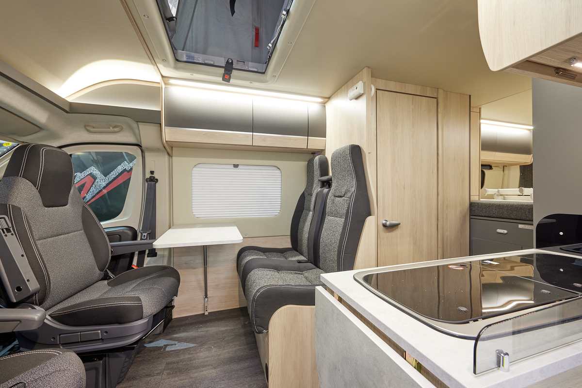 Auto-Trail Expedition 68 has two tone upholstery, with pale wooden furniture.  A hob with extendible flap for additional kitchen work surface is in the kitchen.  The rising roof is open.  The floor is dark wood in colour.