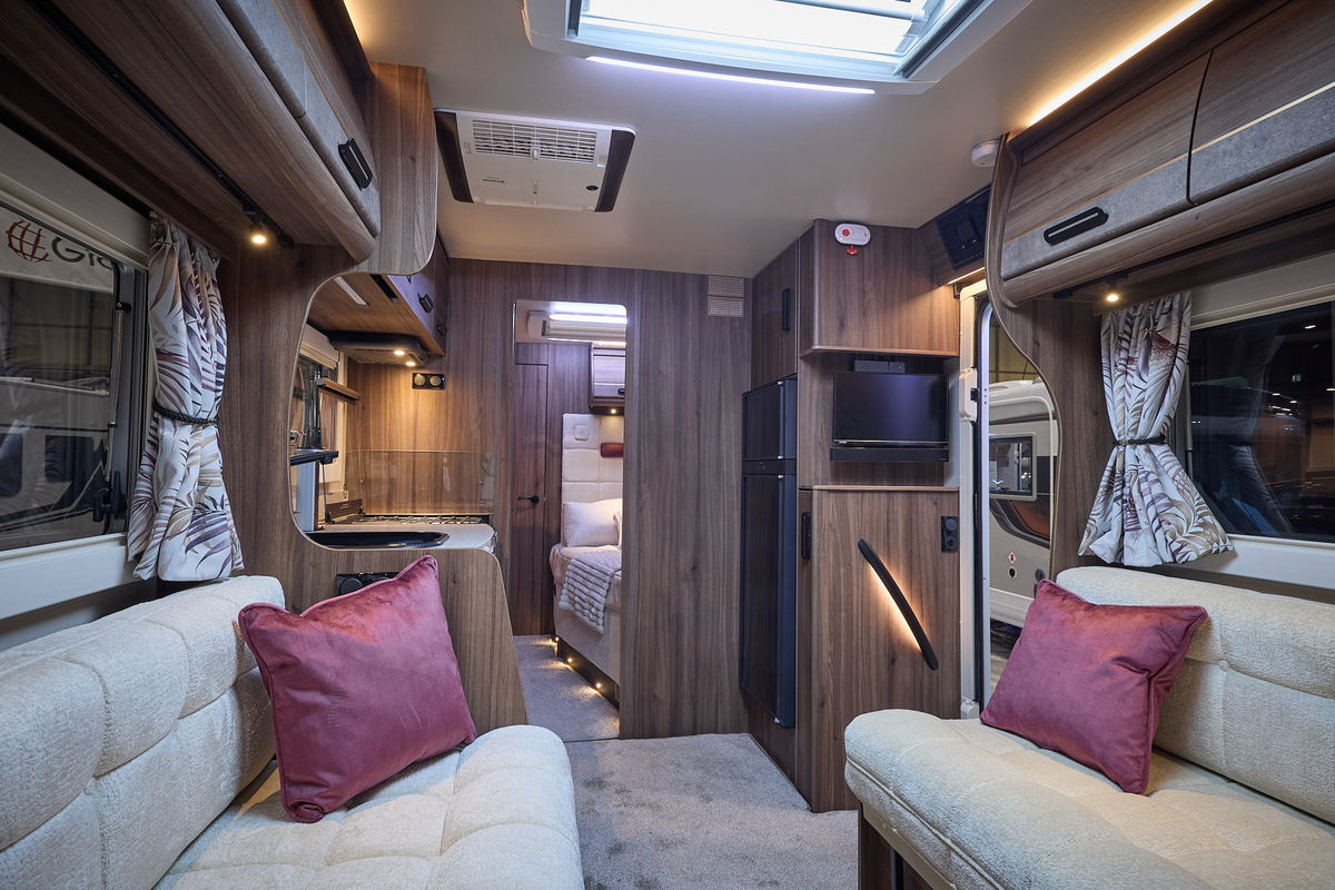 The Auto-Sleeper Burford has a pale grey carpet, with dark wood cabinets.  There is a roof light.  The upholstery is cream with two burgundy cushions.  There is a fixed bed to the rear.