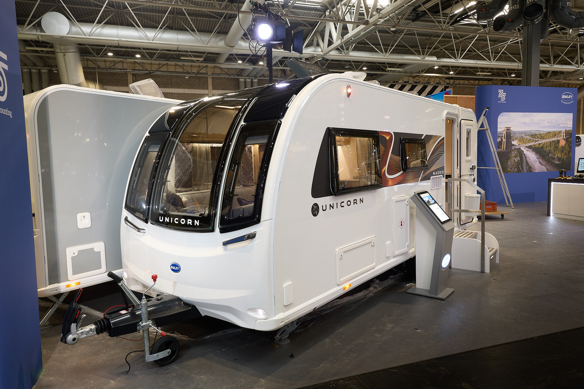 Bailey Unicorn Madrid exterior’s white with a large front window.  The entrance door is open and there are fixed metal steps into its interior.  There is an interactive stand next to the steps.