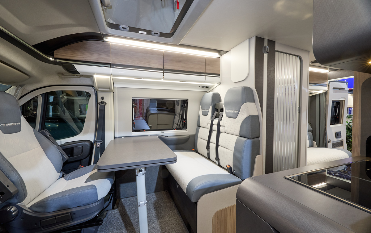 The Swift Carrera 244 has a grey floor and its upholstery is pale grey and blue.  The cabinets are wood with white handles.  The rising roof is open. Towards the rear there is lounge and one of its rear doors are open.