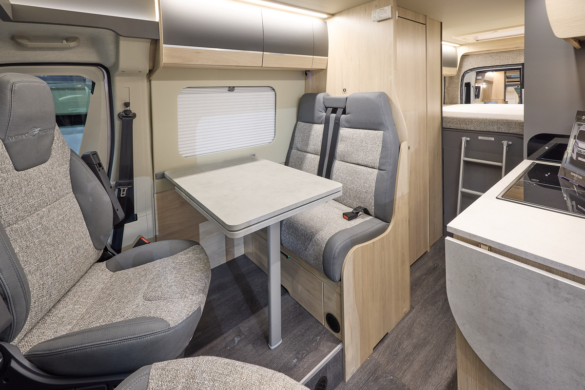 The Auto-Trail Expedition 68 has grey flooring, with pale wood cabinets with pale wood and grey overhead lockers.  The upholstery is grey and cream.  There is a ladder leading to the rear fixed bed.