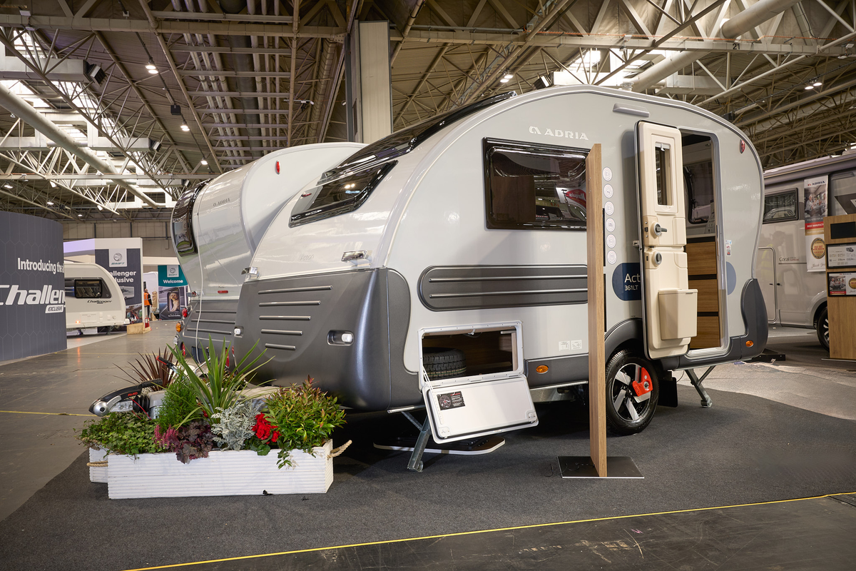 Adria Action exterior, white with dark grey bumpers and trim.  The locker door and main door are open and there are flowers around its hitch in white planters.   