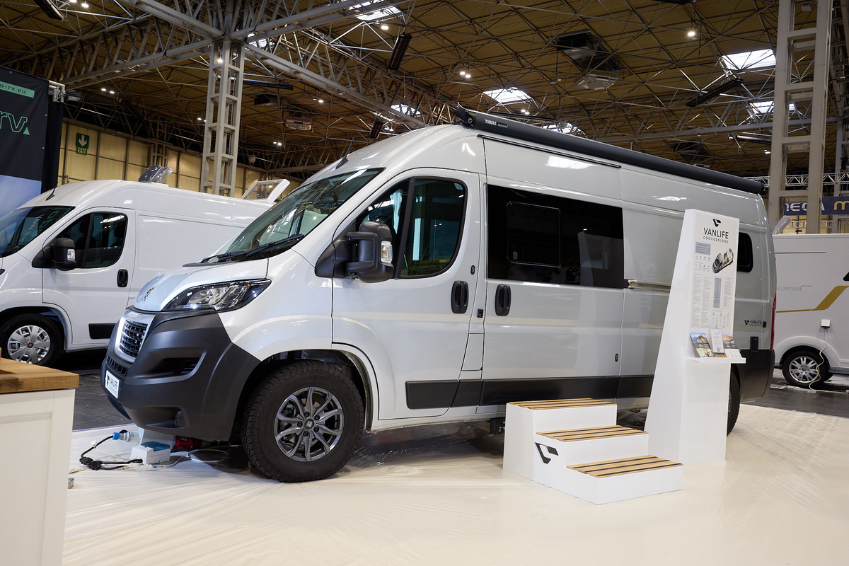 Vanlife Conversions Kunu is silver with black trim.  There are three steps outside the closing sliding door  There is an infomation stand next to the steps.